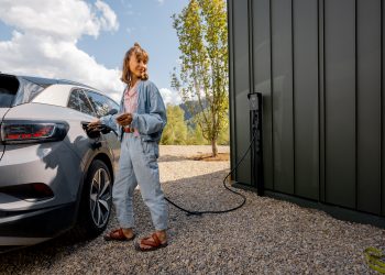 Young,Woman,Plugs,A,Charger,Into,Electric,Vehicle,Standing,With
